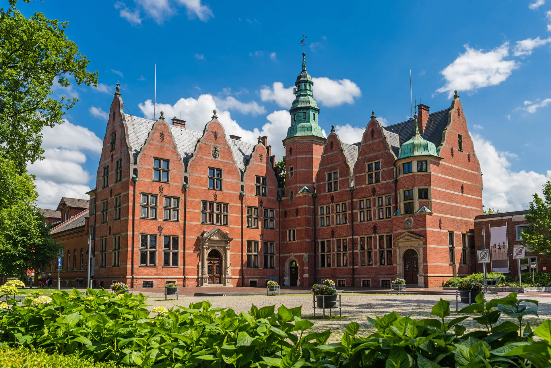 Historisches Backsteingebäude in Aurich mit Turm und grünen Kuppeln, vor blauem Himmel mit Wolken.