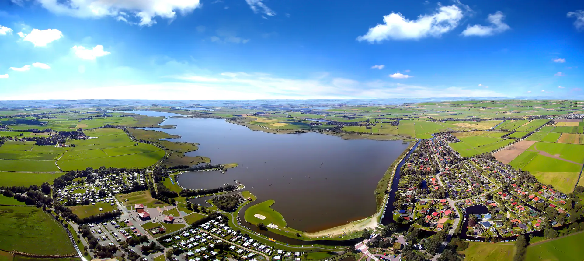 Luftaufnahme von Aurich-Emden: Großer See, Campingplatz, Wohngebiet und grüne Felder unter blauem Himmel.