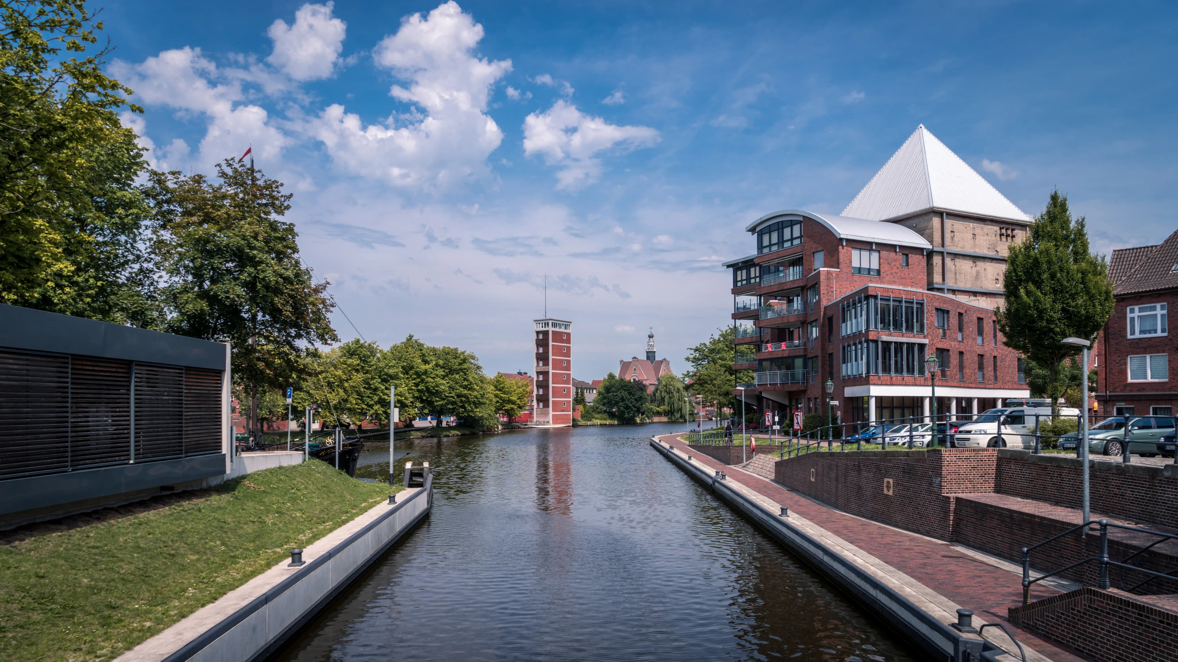 Kanal mit modernen Gebäuden, rotem Turm und grünen Bäumen unter blauem Himmel mit Wolken.